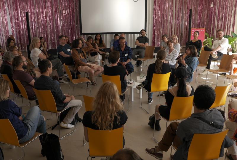 People sitting in a circle of chairs during a moderated group discussion in a bright room, with a projection screen and a pink curtain in the background.