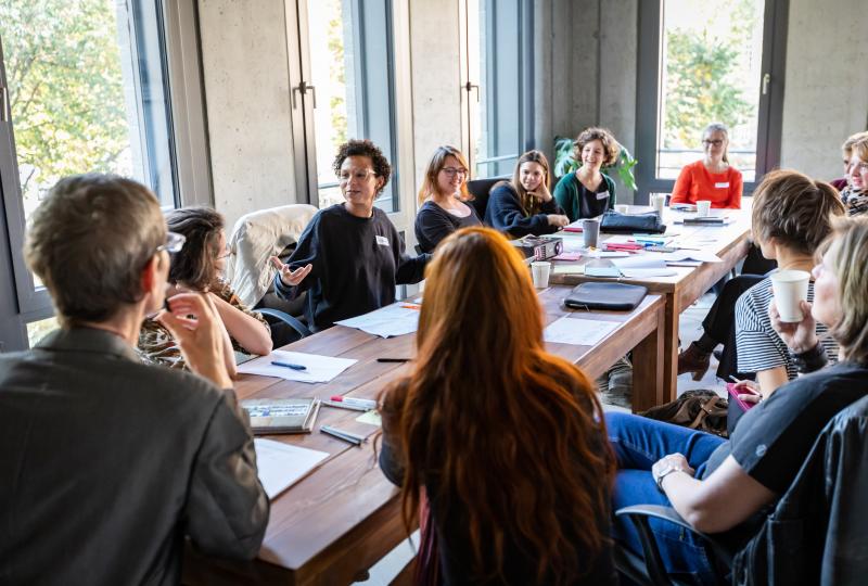 Eine Gruppe von Menschen sitzt an einem langen Besprechungstisch in einem hellen Raum und diskutiert gemeinsam. Auf dem Tisch liegen Unterlagen, Notizblöcke und Getränke. Mehrere Personen hören aufmerksam zu, während eine Person am Tisch spricht.