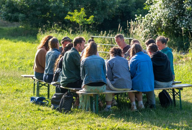 Eine Gruppe von Menschen sitzt mit dem Rücken zum Betrachter auf Bänken. Diese stehen auf einer Wiese im Freien.